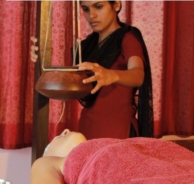 Ayurveda 1 Stehende Frau mit rotem Kleid platzier eine Schale über einer liegenden Frau, die mit einem Handtuch abgedeckt ist